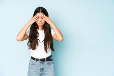 Mavi arka plandaki genç Hintli kadın başı ağrıyor, yüzüne dokunuyor..