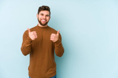Mavi arka planda izole edilmiş genç beyaz adam gülümsüyor ve başparmağını kaldırıyor.