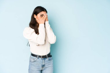 Mavi arka planda izole edilmiş genç Hintli kadın parmaklarının arasından korkmuş ve gergin bir şekilde göz kırpıyor..