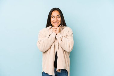 Mavi arka planda izole edilmiş genç bir kadın ellerini çenesinin altında tutuyor ve mutlu bir şekilde yan tarafa bakıyor..