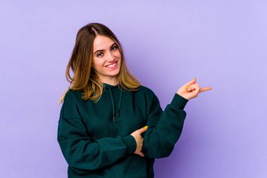 Mor arka planda izole edilmiş genç beyaz kadın işaret parmağıyla işaret ederek gülümsüyor..