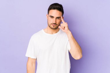 Mor arka planda izole edilmiş genç beyaz bir adam şakağı parmağıyla işaret ediyor, düşünüyor ve bir göreve odaklanıyor..