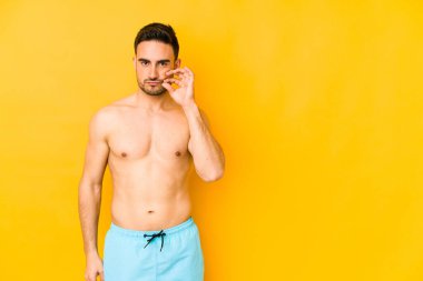 Sarı arka planda mayosu olan genç, beyaz bir adam. Beyaz tenli, dudaklarında saklayan genç bir adam..