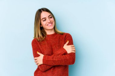 Mavi arka planda izole edilmiş genç beyaz kadın kollarını kavuşturarak gülümsüyor..