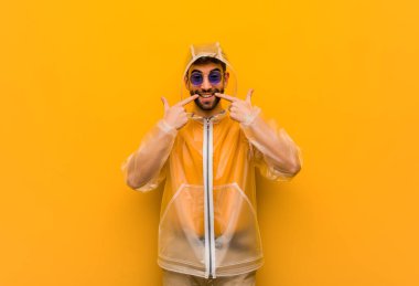 Yağmurluk giyen genç adam gülümsüyor, ağzını işaret ediyor.