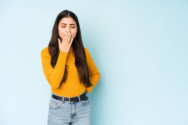 Mavi arka planda izole edilmiş genç bir Hintli kadın esniyor. Ağzı eliyle kapatan yorgun bir hareket sergiliyor..