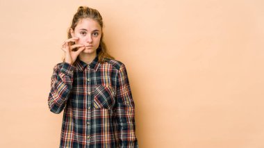 Beyaz tenli genç bir kadın bej arka planda izole edilmiş ve parmakları dudaklarında bir sır saklıyor..