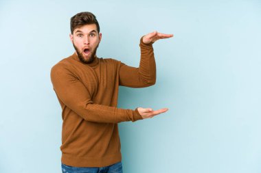 Mavi arka planda izole genç beyaz adam şok ve hayret içinde elleri arasında bir kopya boşluk tutuyor.