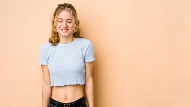 Beyaz tenli genç bir kadın bej arka planda izole edilmiş. Kahkahalar atıyor, gözlerini kapatıyor, rahatlıyor ve mutlu oluyor..