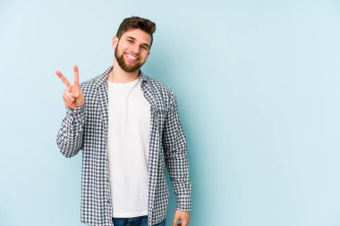 Mavi arka planda izole edilmiş genç beyaz adam zafer işareti gösteriyor ve gülümsüyor..