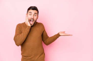 Pembe arka planı olan genç beyaz bir adam avuçlarında fotokopi alanı tutuyor, yanaklarını yana tutuyor. Şaşırmış ve mutlu..