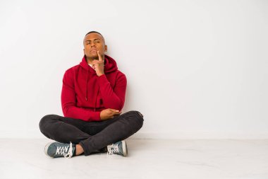 Yerde tek başına oturan genç latin adam şüpheli ve şüpheci bir ifadeyle yana bakıyor..