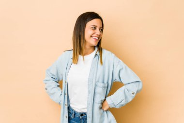 Bej arka planda izole edilmiş genç bir kadın mutlu bir şekilde gülüyor ve ellerini karnının üstünde tutarak eğleniyor..