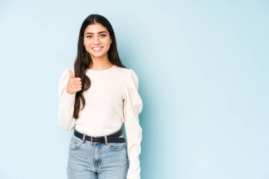 Mavi arka planda izole edilmiş genç Hintli kadın gülümsüyor ve başparmağını kaldırıyor.