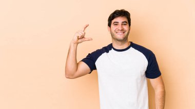 Bej arka planda izole edilmiş genç bir adam işaret parmaklarıyla küçük bir şey tutuyor, gülümsüyor ve kendinden emin..