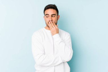 Mavi arka planda izole edilmiş genç beyaz adam esniyor. Ağzı eliyle kapatan yorgun bir hareket sergiliyor..