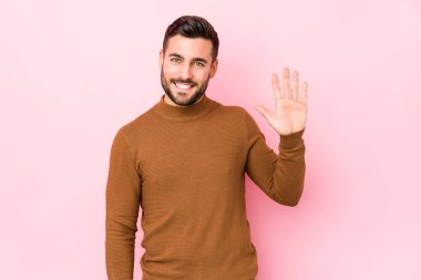 Pembe arka planda genç beyaz bir adam izole edilmiş gülümseyen beş numarayı parmaklarıyla gösteriyor..