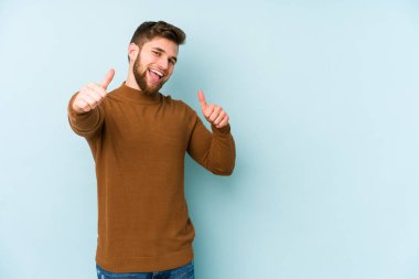 Kafkasyalı genç adam mavi arka planda izole edilmiş. Baş parmağını kaldırıyor, gülümsüyor ve kendinden emin..