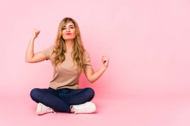 Pembe bir stüdyoda oturmuş, özel bir günü kutlayan genç sarışın beyaz kadın, atlıyor ve enerjiyle kollarını kaldırıyor..