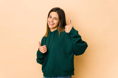 Bej arka planda izole edilmiş genç bir kadın. Baş parmağını kaldırıyor, gülümsüyor ve kendinden emin..