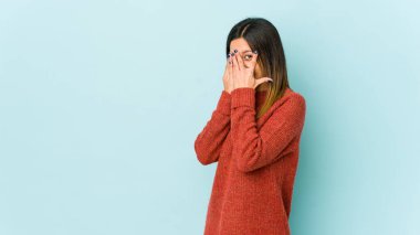 Mavi arka planda izole edilmiş genç bir kadın parmaklarının arasından korkmuş ve gergin bir şekilde göz kırpıyor..