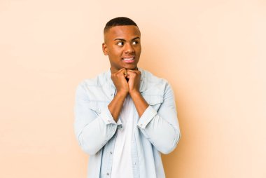 Bej arka planda izole edilmiş genç latin adam ellerini çenesinin altında tutuyor, mutlu bir şekilde yana bakıyor..