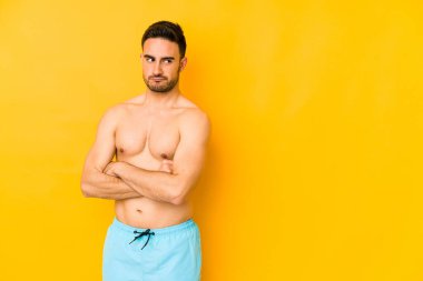 Sarı arka planda mayosu olan genç, beyaz bir adam. Mutsuz, alaycı bir ifadeyle kameraya bakan beyaz bir genç..