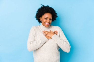 Orta yaşlı Afro-Amerikalı bir kadın mavi arka planda izole edilmiş, dost bir ifadeye sahip, avuçlarını göğsüne bastırıyor. Aşk konsepti.