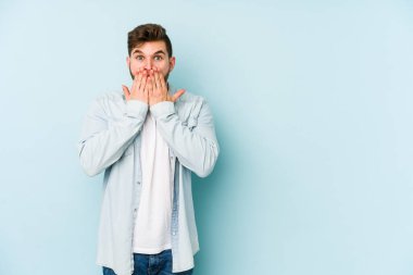 Mavi arka planda izole edilmiş genç beyaz adam şok olmuş, ağzı ellerle kaplanmış, yeni bir şey keşfetmeye hevesli..