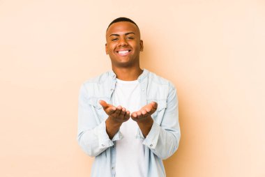 Genç Latin bir adam bej arka planda izole edilmiş. Elinde avuç içi olan bir şey var ve kameraya sunuyor..