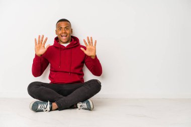 Yerde tek başına oturan genç latin adam on numarayı elleriyle gösteriyor..