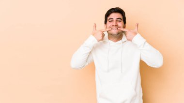 Bej arka planda izole edilmiş genç adam gülümsüyor, parmaklarını ağzına doğrultuyor..