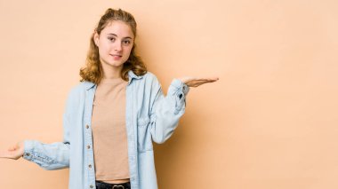 Beyaz tenli genç bir kadın, bej arka planda izole edilmiş. Kafası karışmış ve omuz silkiyor. Kopya alanını tutmak için..