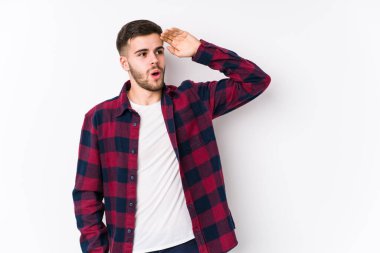 Beyaz arka planda poz veren beyaz tenli genç bir adam çok uzaklara bakıyor ve elini alnına koyuyor..