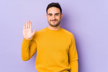Mor arka planda izole edilmiş genç beyaz adam gülümseyerek beş numarayı parmaklarıyla gösteriyor..