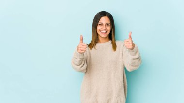 Mavi arka planda izole edilmiş genç bir kadın gülümsüyor ve baş parmağını kaldırıyor.