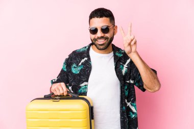Elinde bir bavul tutan genç latin adam zafer işareti gösteriyor ve gülümsüyor..