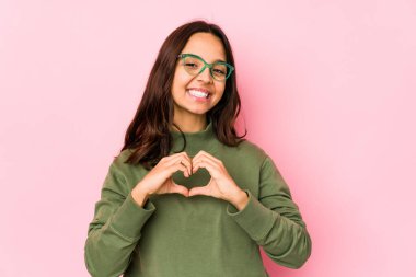 Latin kökenli genç bir kadın izole edilmiş gülümsüyor ve elleriyle kalp şeklinde bir şey gösteriyor..