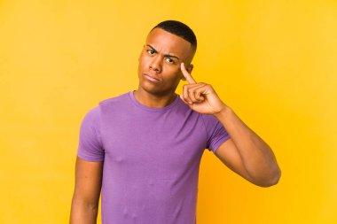 Sarı arka planda izole edilmiş genç Latin bir adam şakağı parmağıyla işaret ediyor, düşünüyor ve bir göreve odaklanıyor..