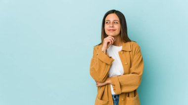 Genç bir kadın mavi arka planda izole edilmiş düşünüyor, yukarı bakıyor, yansıtıyor, düşünüyor, bir fantezi kuruyor..