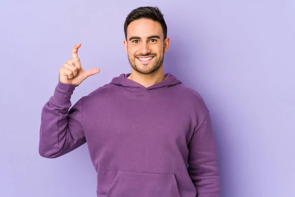 Young caucasian man isolated on purple background holding something little with forefingers, smiling and confident.