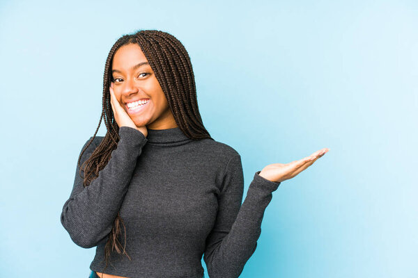 Young african american woman isolated on blue background holds copy space on a palm, keep hand over cheek. Amazed and delighted.