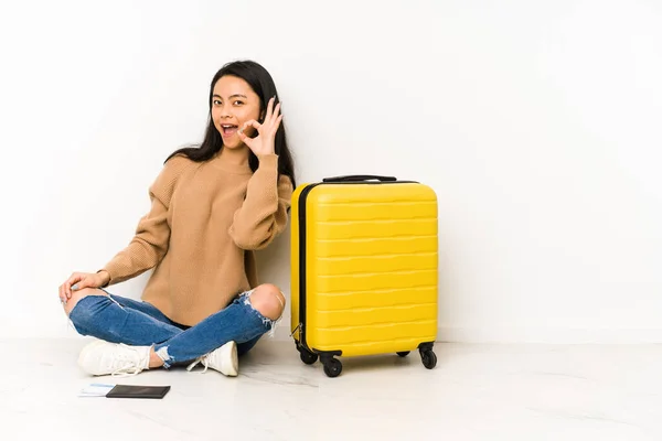 若いです中国の旅行者女性座っています上の床にスーツケース隔離されたウィンクA目と保持しています手で正しいジェスチャー — ストック写真