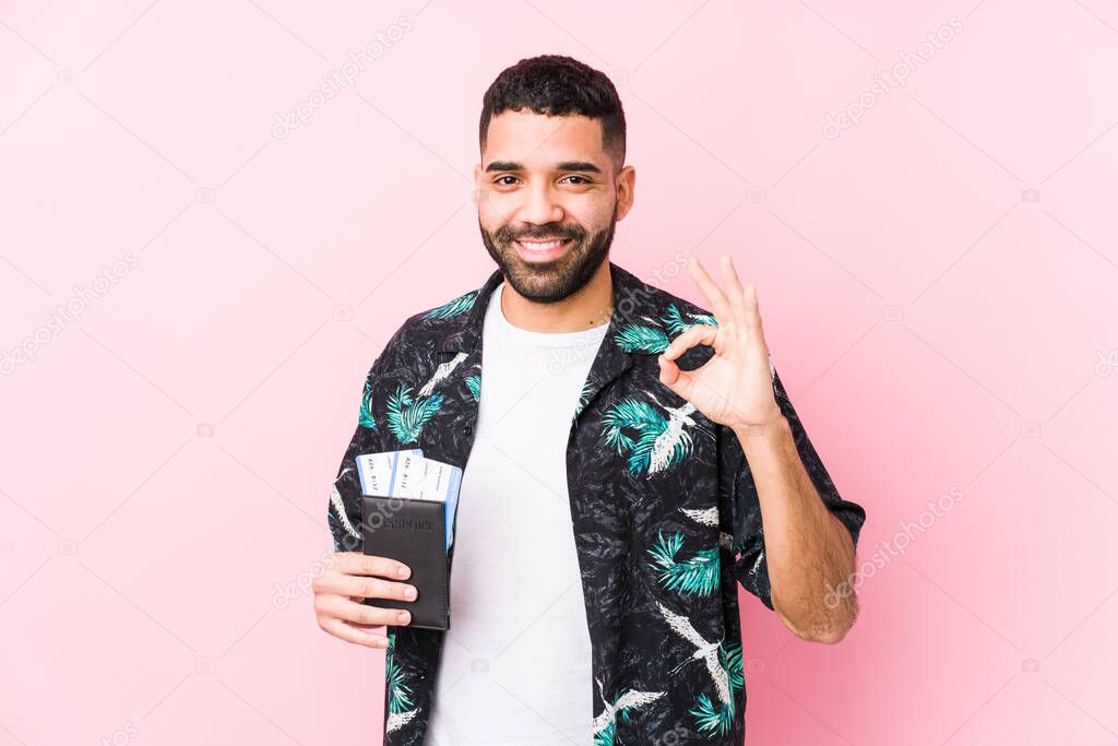 Young arabian cool man holding a boarding passes isolated cheerful and confident showing ok gesture.