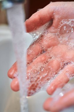 Coronavirus önleyici sabunla yıkanan eller açık musluk ve önde akan su ile beyaz lavaboda.