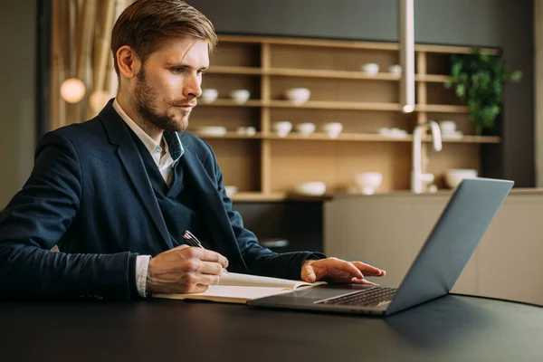 Confident businessman working remotely from the home kitchen with a laptop and notebook. Side view of a young man in smart casual wear making notes during zoom video meeting conference call remote