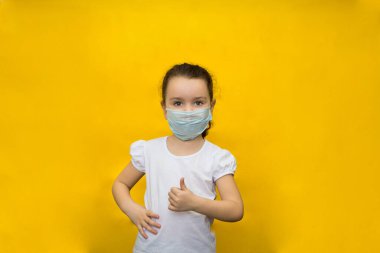 Koruyucu maskeli küçük bir kız sarı arka planda baş parmağının yukarı kaldırma hareketini gösteriyor. Koronavirüse karşı koruma.