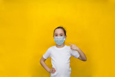 Beyaz tişörtlü ve koruyucu maskeli küçük kız parmağını maskeye doğrultuyor. Koronavirüse karşı koruma.