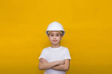 İnşaat kaskı takmış gülümseyen küçük bir kız sarı arka planda kollarını göğsünde kavuşturdu..
