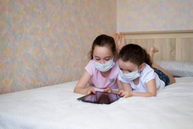 Koruyucu maskeli küçük kızlar tablete bakarlar. Evde, yatakta kendi kendini izole ederek uzaktan eğitim öğrenme.